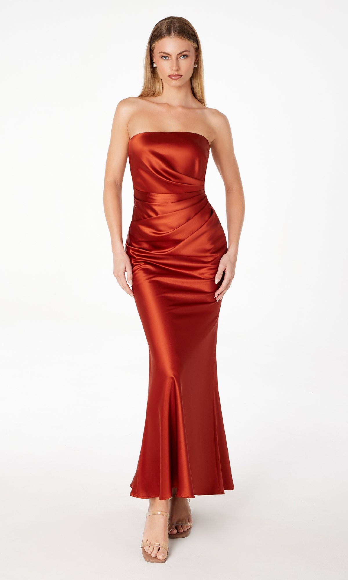 A woman poses against a plain background wearing the Astee T2006 Strapless Long Satin Maxi Prom Dress in shiny rust-orange with a fitted silhouette and mermaid hem, paired with nude high-heeled sandals.